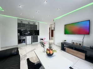 Una sala de estar moderna con un televisor de pantalla plana, un sofá de cuero negro, una mesa de centro con fruta, y una cocina al fondo.