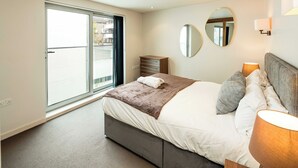 Une chambre à coucher comprenant un lit, des lampes de chevet, une commode, un miroir et une porte-fenêtre coulissante donnant sur l’extérieur.