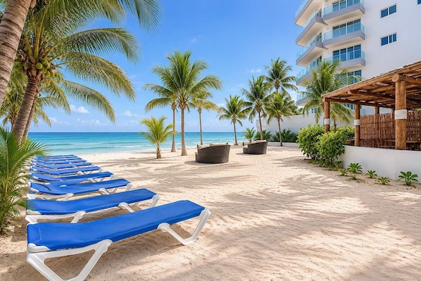 Une plage avec des chaises longues bleues, des palmiers et un bâtiment doté de balcons.