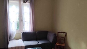 A room with a sofa, a wooden table, a chair, a hanging light fixture, and a window with curtains. - 2-room apartment in a large, quiet house with garden and shared spaces (Vence)