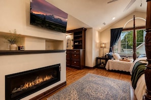 A bedroom with a fireplace, a bed with a canopy, a nightstand, a window with curtains, and a mounted TV.