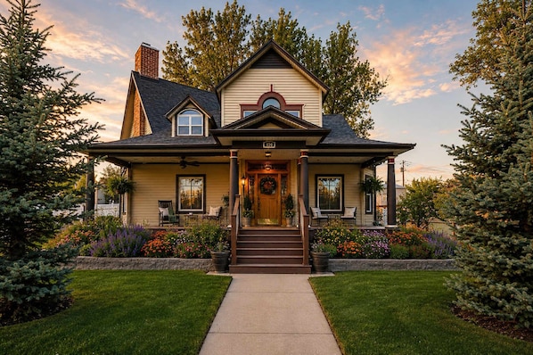 A two-story house with a front porch, a chimney, and a front yard with flowers and trees.