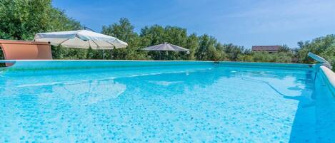 Una piscina azul cristalina con fondo de azulejos, rodeada de árboles y un cielo despejado.
