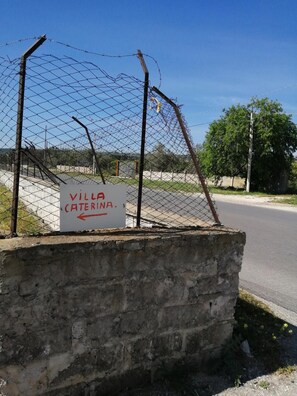 A sign for 'Villa Caterina' with a directional arrow, a chain-link fence, and a road in the background.