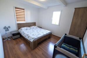 A bedroom with a wooden bed, a bedside fan, a small nightstand, a window with blinds, and a wooden wardrobe.