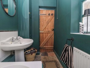 Une salle de bain avec un lavabo blanc, une porte en bois, une fenêtre avec une lampe noire et une serviette à rayures accrochée à un radiateur.