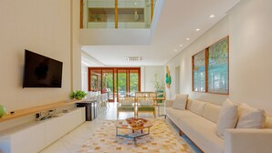Ein modernes Wohnzimmer mit einer Couch, einem Couchtisch und einem Fernseher. Ein Balkon mit Glasgeländern und Blick auf Grünflächen.