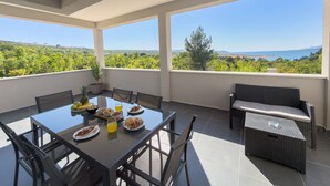 A balcony with a table set for breakfast, a sofa, and a view of a green landscape. - Apartment Temida with sea view (Rovanjska)