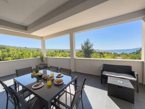 A balcony with a table set for breakfast, a sofa, and a view of a green landscape. - Apartment Temida with sea view (Rovanjska)