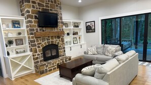 A living room with a stone fireplace, a television mounted on the wall, a grey sofa, and a wooden floor. - Stunning gated ranch w/ breathtaking views and modern amenities, on 103 acres. (Sand Springs)