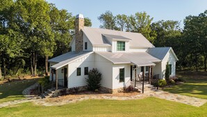 A two-story house with a stone chimney, a covered porch, and a stone pathway leading to the entrance. - Stunning gated ranch w/ breathtaking views and modern amenities, on 103 acres. (Sand Springs)