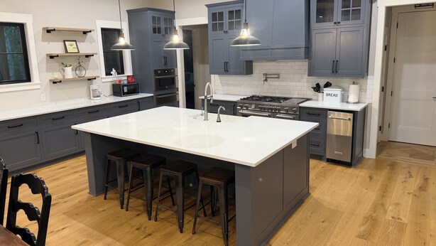 A modern kitchen with grey cabinets, a central island with stools, a stove, and overhead pendant lights. - Stunning gated ranch w/ breathtaking views and modern amenities, on 103 acres. (Sand Springs)