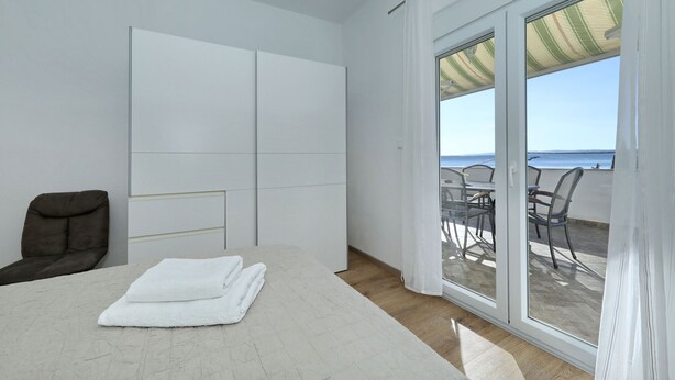Un dormitorio con cama, una silla, armario y puerta corrediza de vidrio que da a una zona de estar exterior con vista al mar.