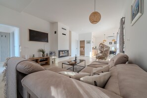 Ein modernes Wohnzimmer mit einem großen beigefarbenen Sofa, einem Couchtisch und einem Fernseher. Im Hintergrund steht ein Korbsessel und ein Essbereich.