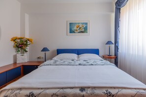 Ein ordentlich bezogenes Bett mit blauem Kopfteil, ein Nachttisch mit Lampe und ein Wandbild mit Blumenmotiven.