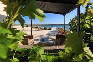 Ein überdachter Außenbereich mit Korbmöbeln, einem Glastisch und Blick auf das Meer.