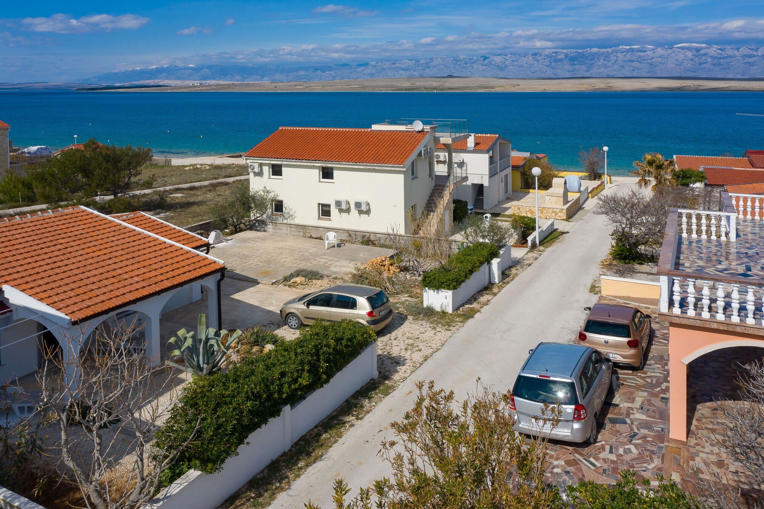 Un quartier résidentiel avec des maisons, une route asphaltée, des voitures garées et, au loin, une vue sur la mer et les montagnes.