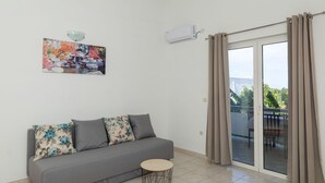 Een woonkamer met een grijze bank, een klein rond tafeltje, een wandairconditioner en een schuifdeur van glas die uitkomt op een buitengebied.