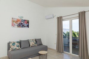 Ein Wohnzimmer mit grauem Sofa, einem kleinen runden Tisch, einer Wand-Klimaanlage und einer Glastür, die zu einem Außenbereich führt.