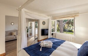 Ein Schlafzimmer mit Bett, Fernseher, Fenster mit Blick auf Bäume, einer Couch und einem Spiegel.