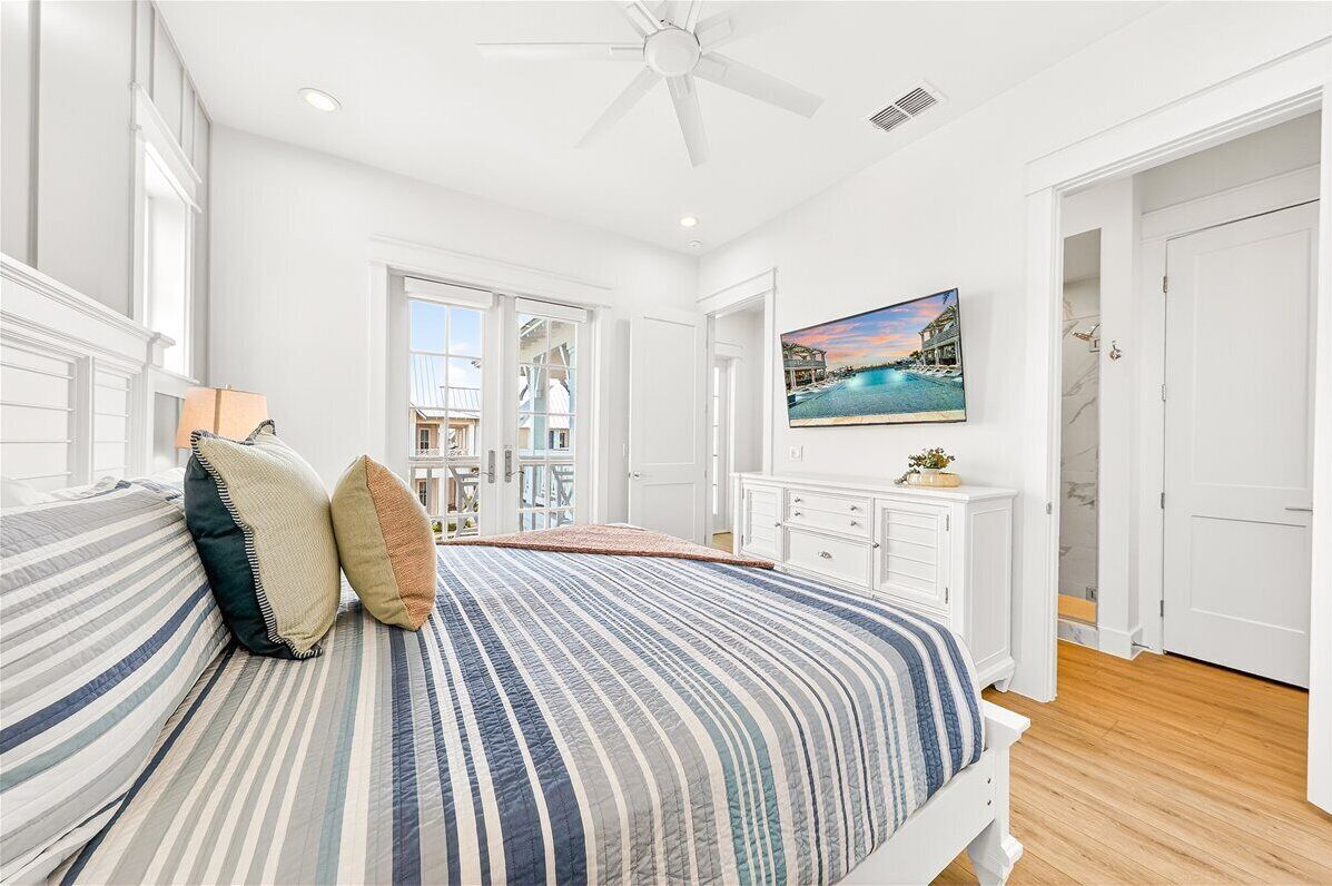 A bedroom with a bed, a ceiling fan, a painting of a pool, and a view of a balcony.