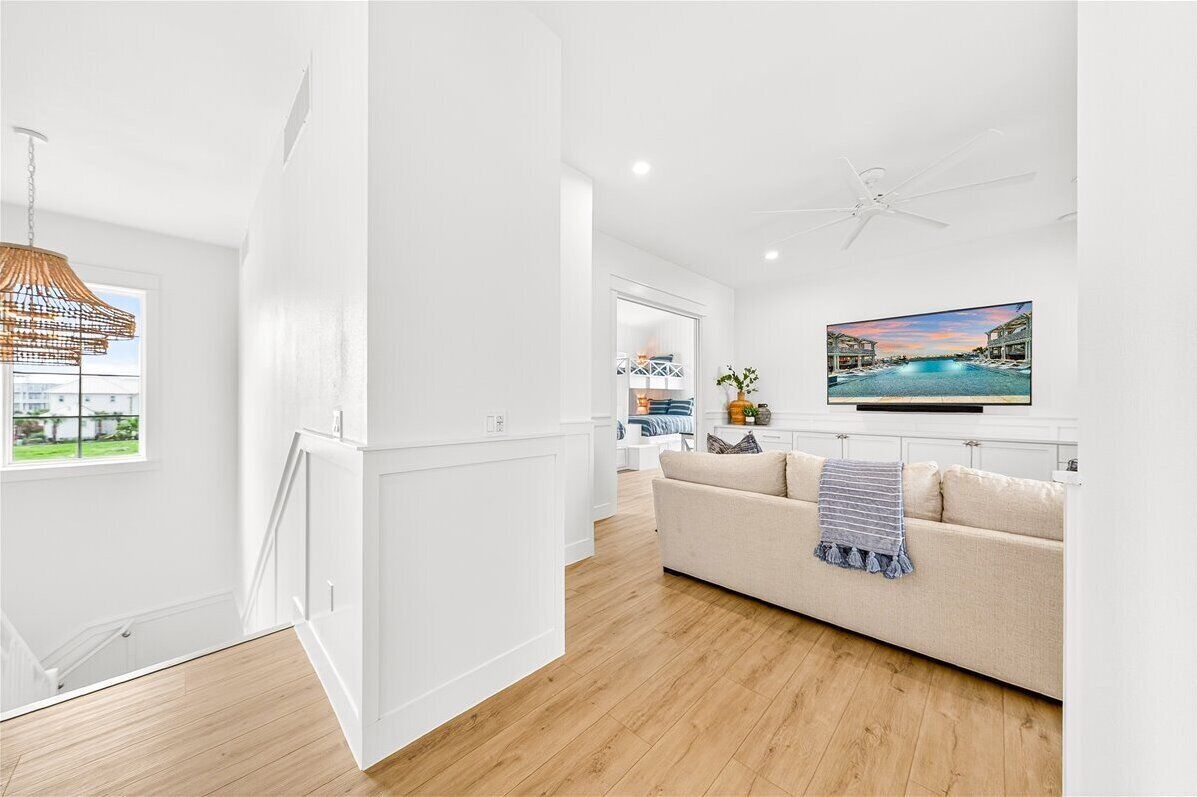 A modern living room with a wooden floor, white walls, a ceiling fan, a TV mounted on the wall, and a window with a view of a pool.