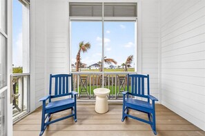 A balcony with two blue chairs, a small white table, and a large window with a view of palm trees and a building. - Toes in the Sand 106 TAI (Port Aransas)