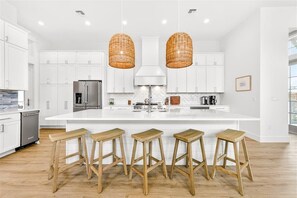 A modern kitchen with white cabinetry, a large island with stools, a stainless steel refrigerator, and two wicker pendant lights. - Toes in the Sand 106 TAI (Port Aransas)