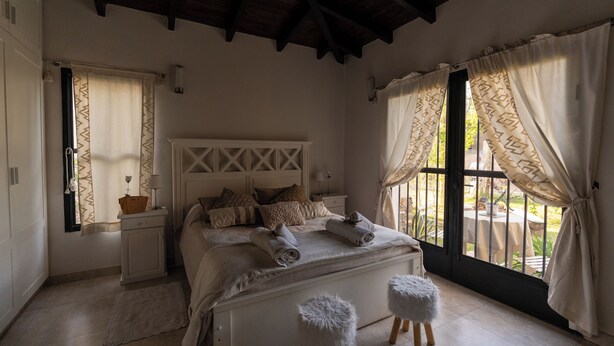 A bedroom with a bed, bedside tables, a window with curtains, and a view of the outdoors. - Casa Andina: Rustic Warmth, Fire Corner and Total Relaxation in Chacras (Luján de Cuyo)
