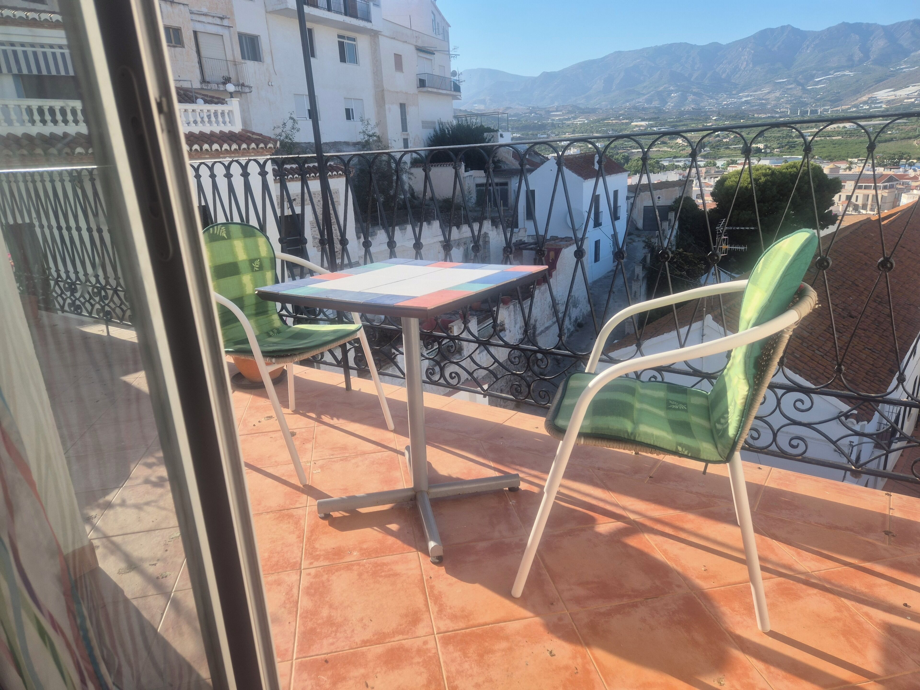 Un balcone con tavolo e sedie, vista su edifici e montagne, e un pavimento piastrellato.