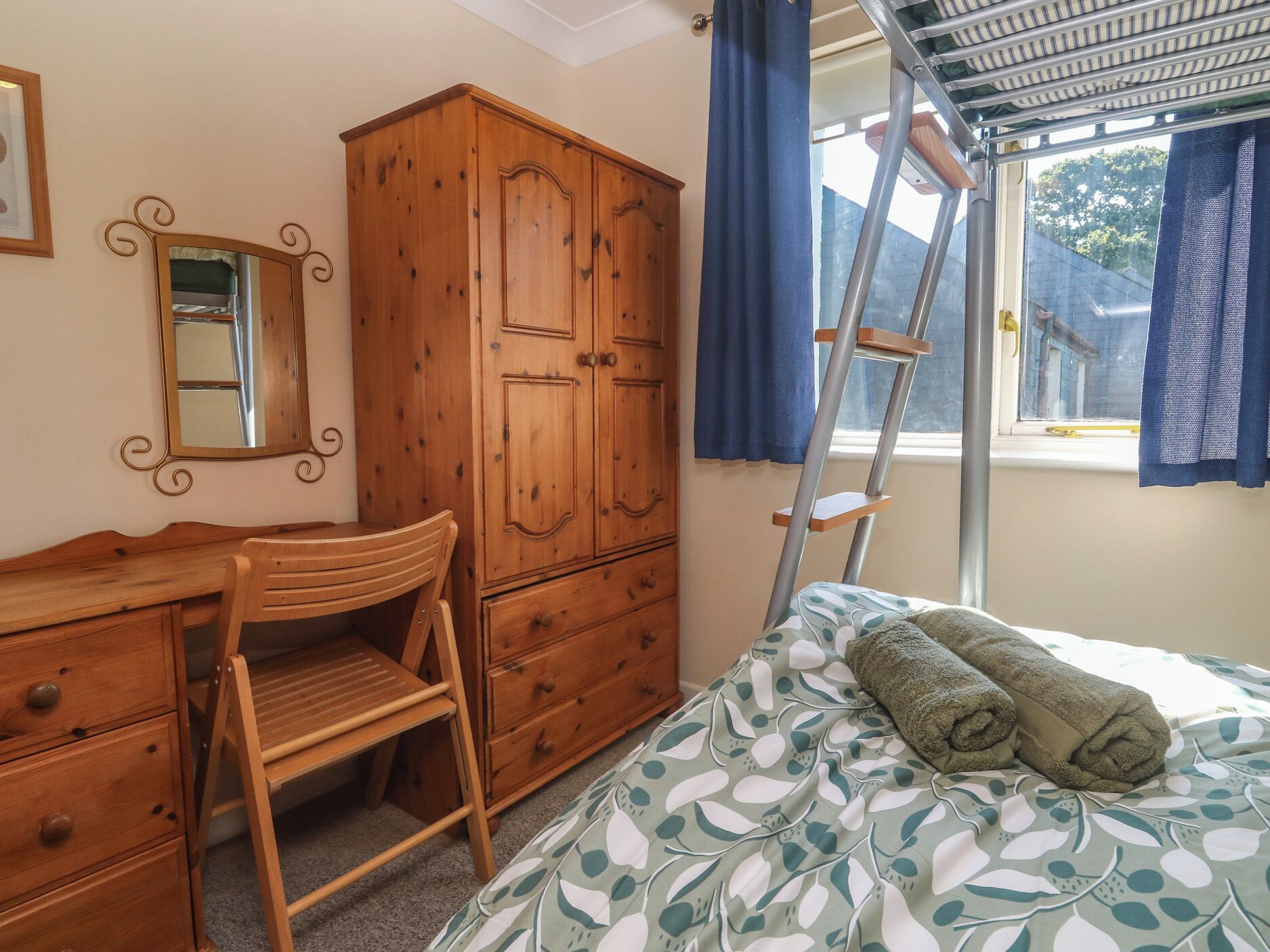 Een slaapkamer met een houten garderobe, een bureau met laden, een stoel, een bed met een beddengoed met dessin, en een raam met gordijnen.