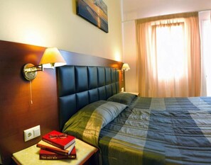 A hotel room with a bed, two bedside lamps, a nightstand with books, and a painting on the wall. - BELVEDERE AEOLIS (Lesvos)