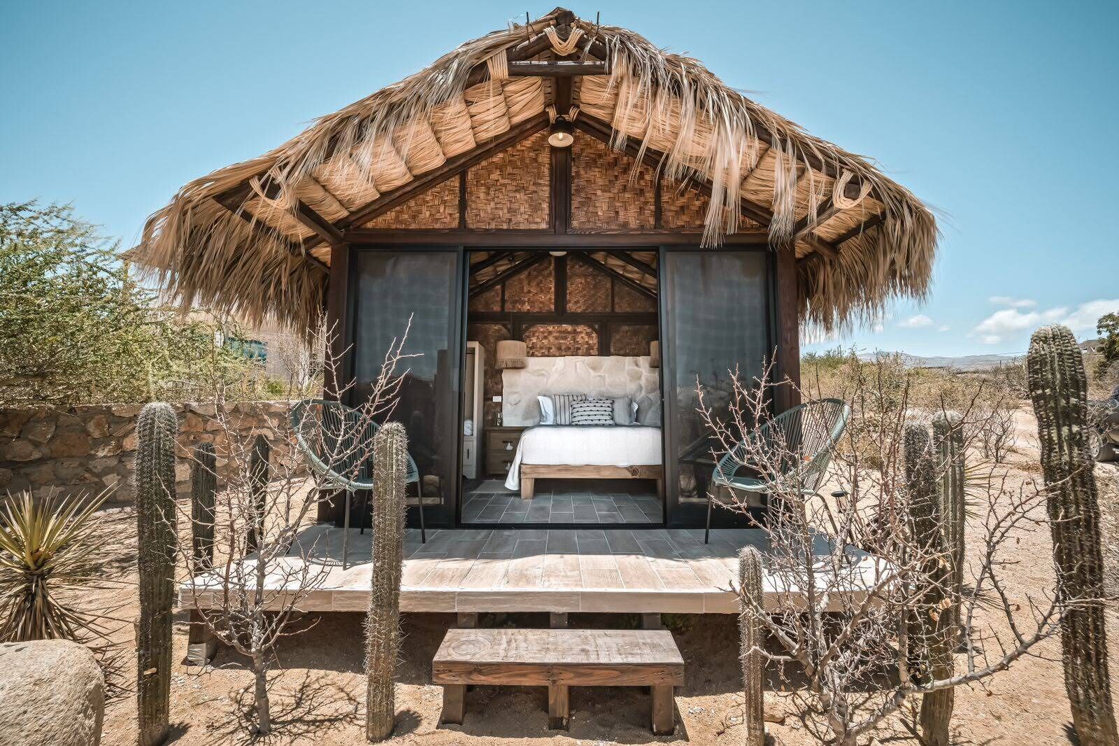 Une structure à toit de chaume comprenant un lit, un banc et des cactus, le tout situé dans un environnement désertique.