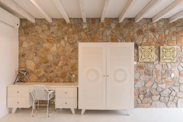 A room with a stone wall, a white wardrobe, a desk with a chair, and decorative wall art.