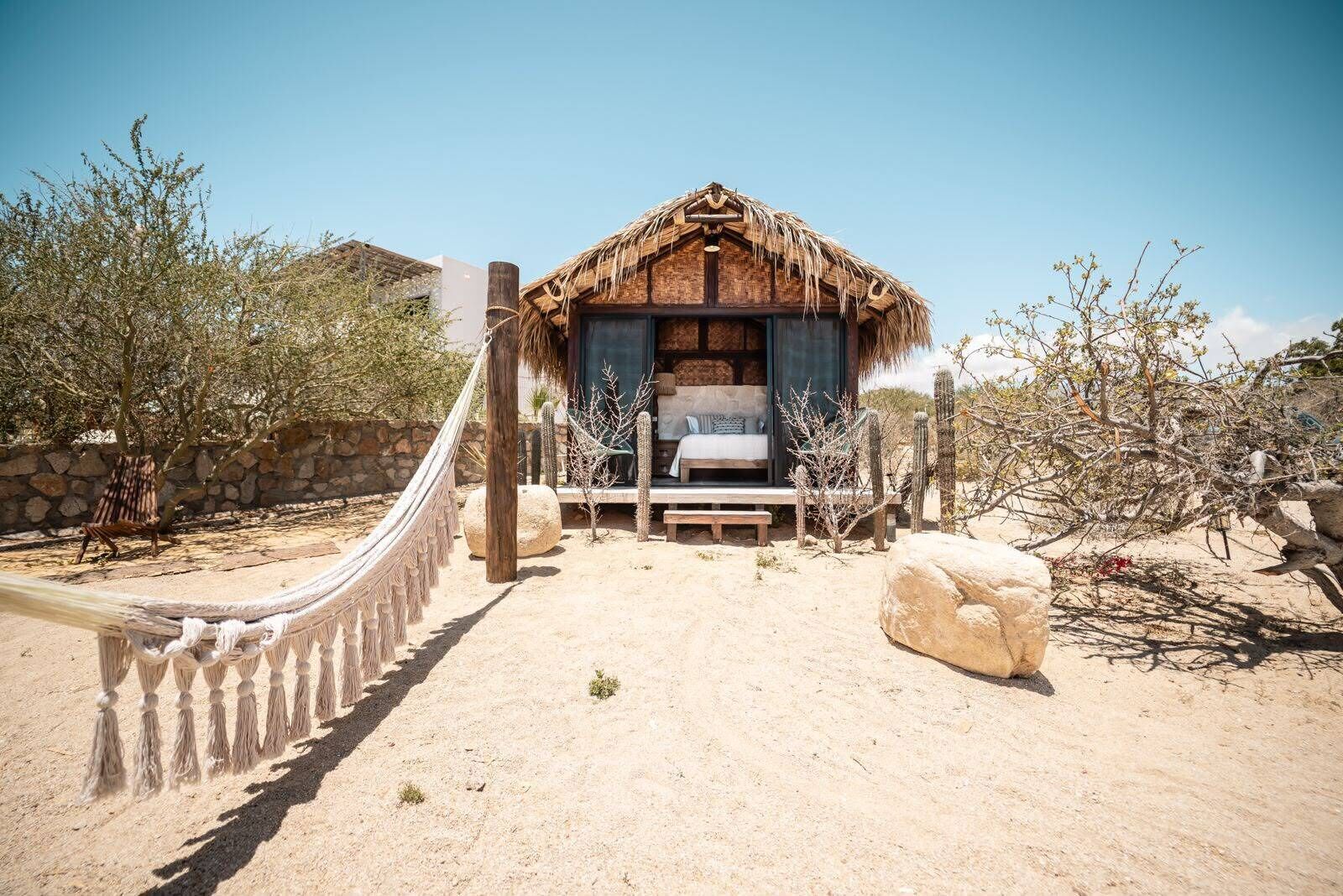 A thatched-roof structure with a bed, a hammock, and a stone wall in a desert setting.