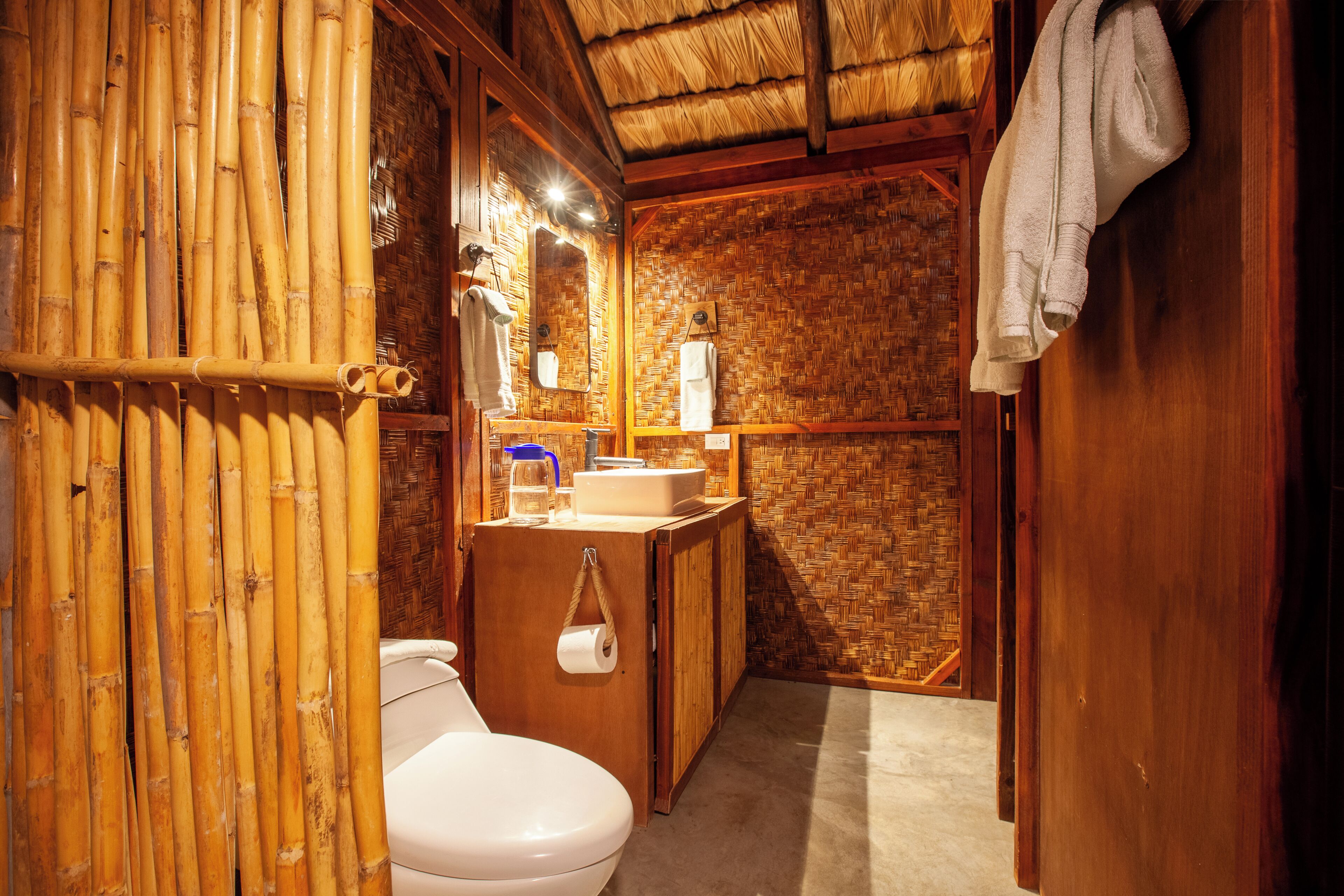 A bathroom with a bamboo wall, a toilet, a sink with a mirror, and a towel hanging on the wall.