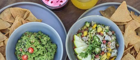 Two bowls of food, one with guacamole and the other with a fruit salad, and two drinks on a table.