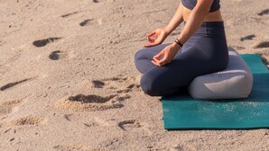 Una persona sentada en una esterilla de yoga sobre una playa arenosa, con huellas en la arena, mirando al océano.