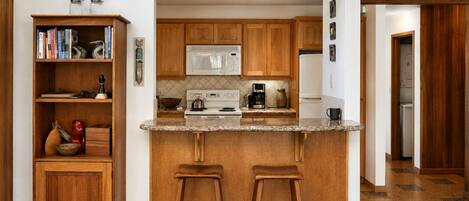A kitchen with wooden cabinets, a granite countertop, a microwave, a stove, and two bar stools.