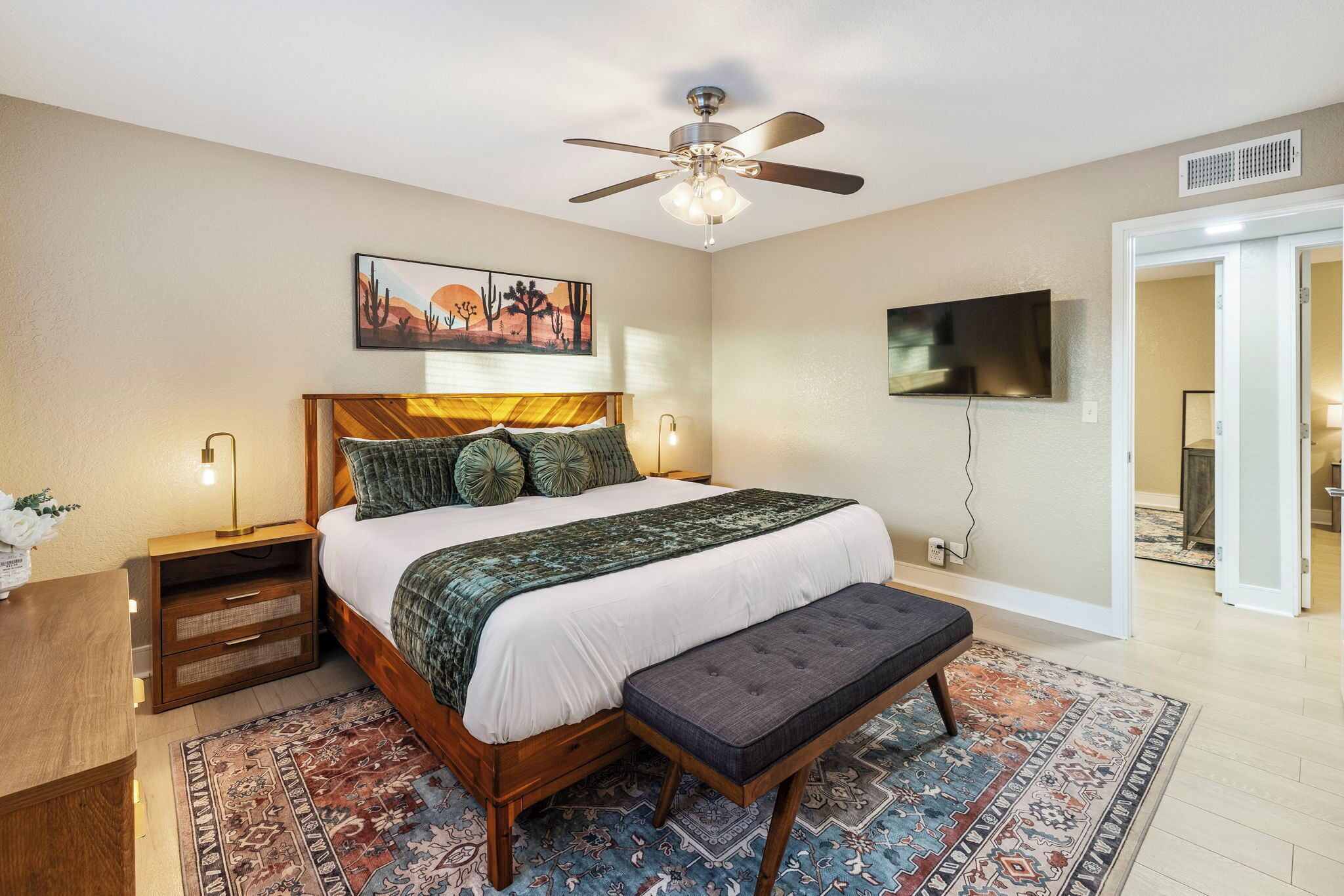 A bedroom with a bed, a bench, a ceiling fan, a TV, and a door leading to another room.