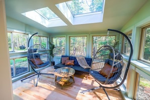 A sunlit room with a sofa, two hanging chairs, a coffee table, and large windows.