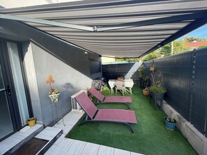 Eine überdachte Terrasse mit Kunstgrasanlage, zwei Lounge-Stühlen, einem Tisch und Topfpflanzen.
