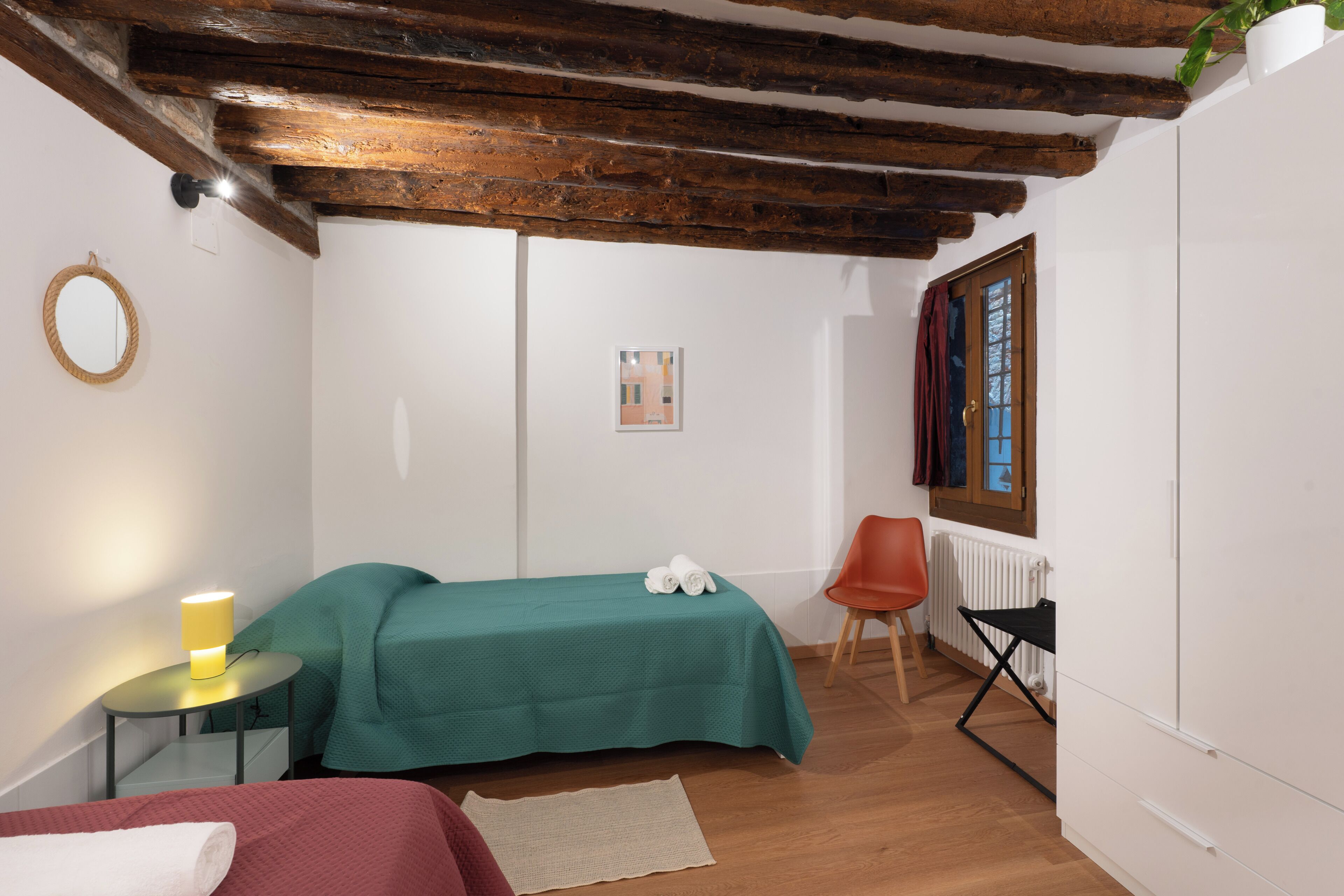 A room with a bed, a chair, a table with a lamp, a radiator, and a window with curtains.