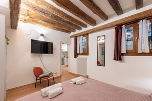 Ein Schlafzimmer mit Holzbalkendecke, einem Flachbildfernseher, einem roten Sessel, einem Bett mit weißen Handtüchern und einem Fenster mit Vorhängen.