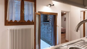 Una cuina amb un vàter d'acer inoxidable, bigues de sostre de fusta, un radiador blanc i una vista per una porta oberta.