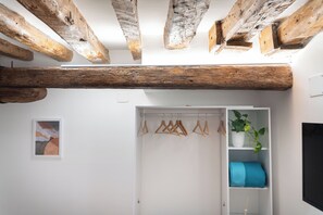 Ein Zimmer mit freiliegenden Holzbalken, einem weißen Schrank mit Kleidung und einem blauen Kissen sowie einer Topfpflanze.
