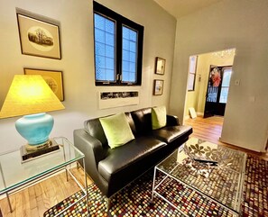 Ein Wohnzimmer mit einem schwarzen Ledersofa, einem Glastisch, einer blauen Lampe, gerahmten Bildern an der Wand und einem Fenster mit mattiertem Glas.