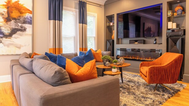 Ein modernes Wohnzimmer mit grauem Sofa, orange- und blauem Kissen, einem Holztisch und einem Kamin.