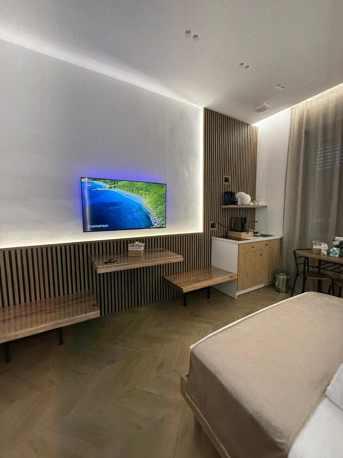 A modern hotel room with a large flat-screen TV mounted on the wall, a wooden bench, a small kitchen area, and a bed.