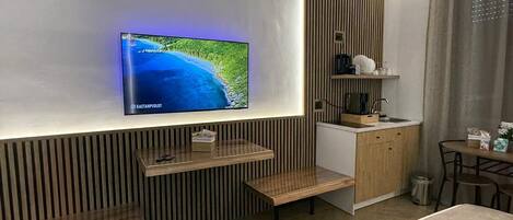 A modern hotel room with a large flat-screen TV mounted on the wall, a wooden bench, a small kitchen area, and a bed.